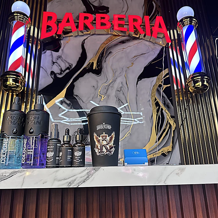 Letrero rojo BARBERIA entre dos postes de barbero iluminados, sobre un muro de mármol blanco con vetas negras y doradas; abajo, una repisa con lociones y un termo negro
