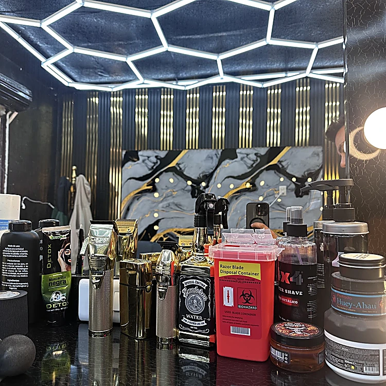 Mostrador negro con máquinas de corte doradas, lociones y ceras; al fondo, el techo de paneles de luz blanca, los listones dorados y el mural de mármol