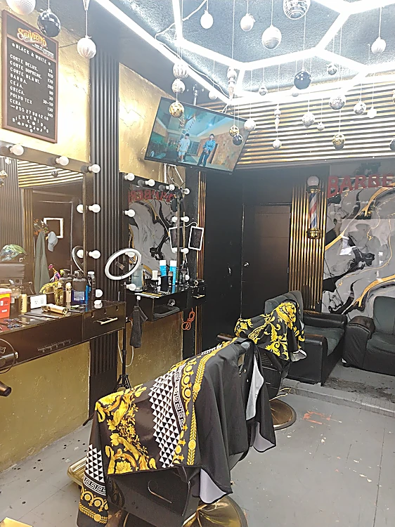 Sillón de barbero cubierto con una capa negra y dorada, frente al espejo con focos y el muro de listones dorados