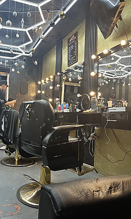 Sillones de barbero negros con base dorada frente a espejos con focos, bajo el techo de paneles de luz